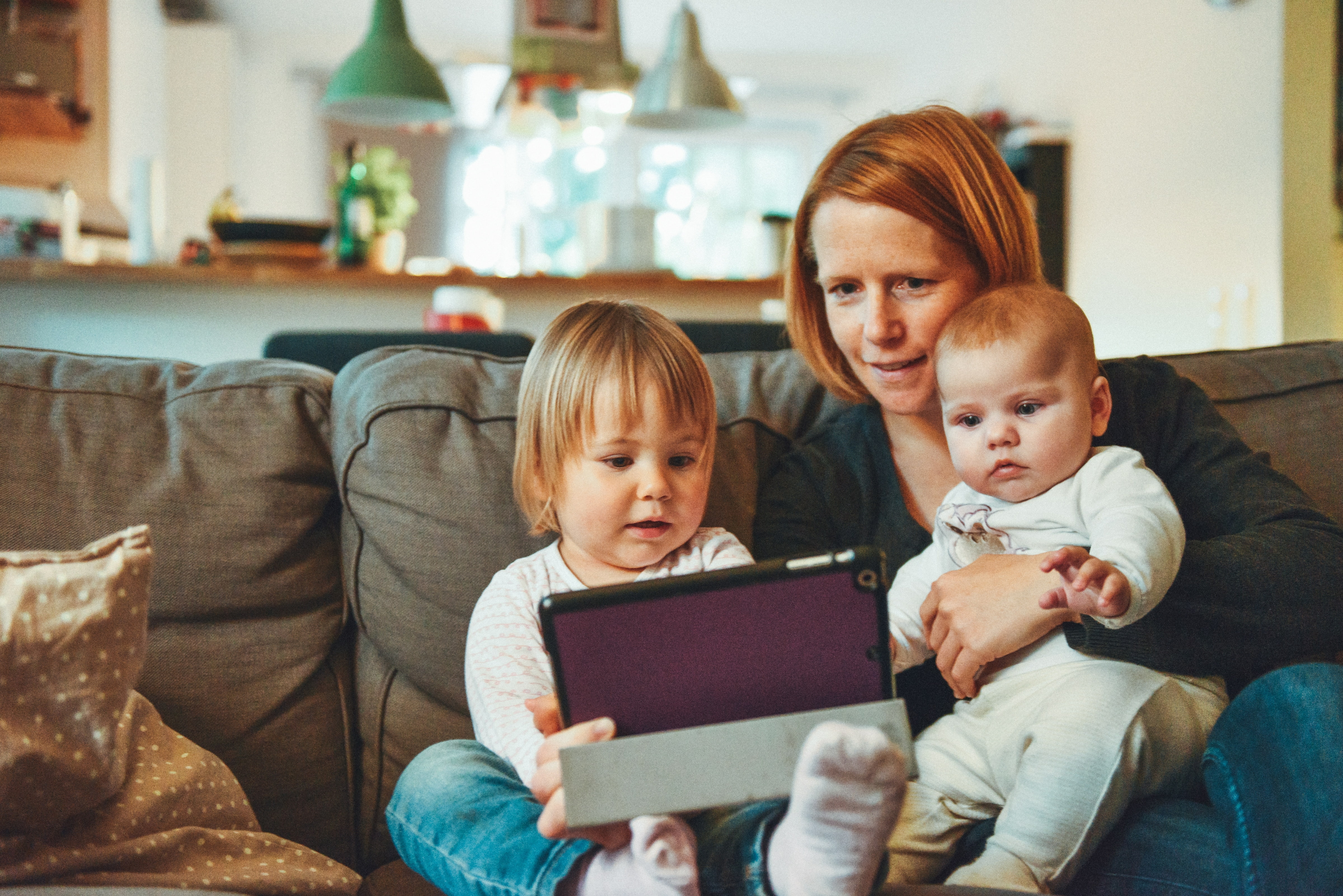 Mom on couch with an infant and a toddler. They are using a tablet together.