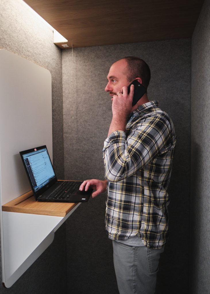 man talks on phone in a makeshift phone booth with laptop stand and laptop open