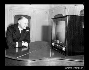 an older man watches an old fashioned tv while leaning on a piano