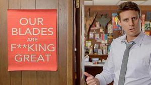 dollar shave club ad with a man pointing to a sign that says "our blades are f**king great"