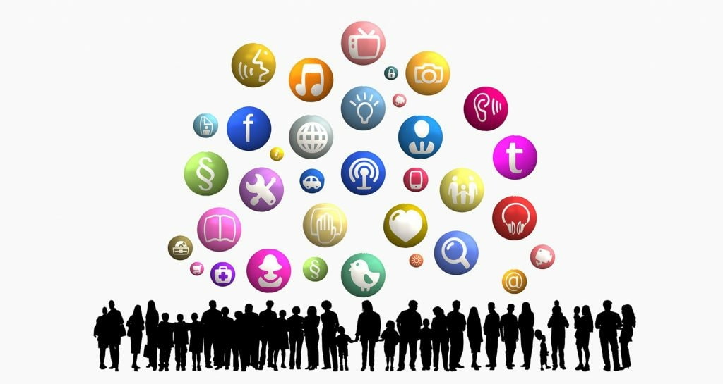 Many standing people in silhouette beneath colorful balloons with icons in them.