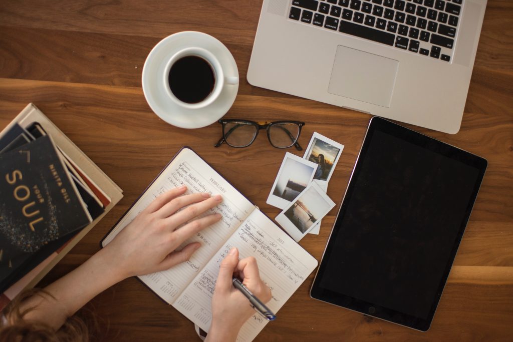 An overhead view of a women sitting at a table near a laptop and writing in a planner.