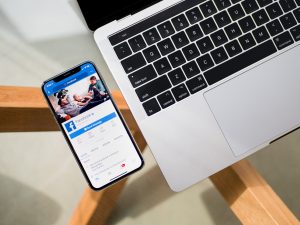 An iPhone and a laptop on a glass table, the phone has Facebook open on the screen.