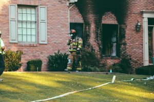 A firefighter, in full gear, walking out of a brick home that was recently on fire.