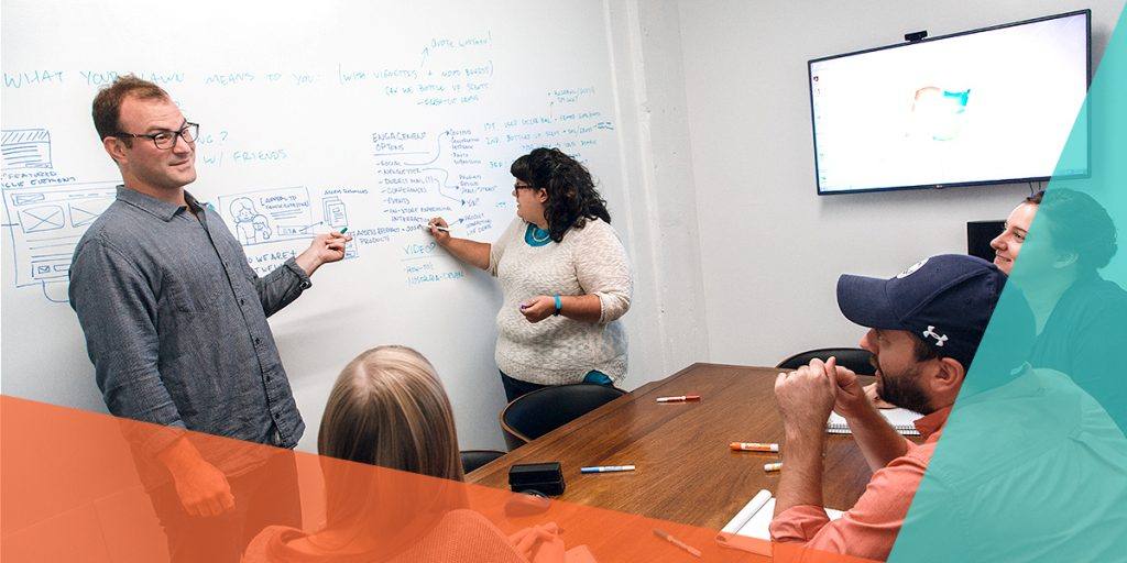 two workers standing at head of conference room writing on the wall talking to two workers sitting at a table