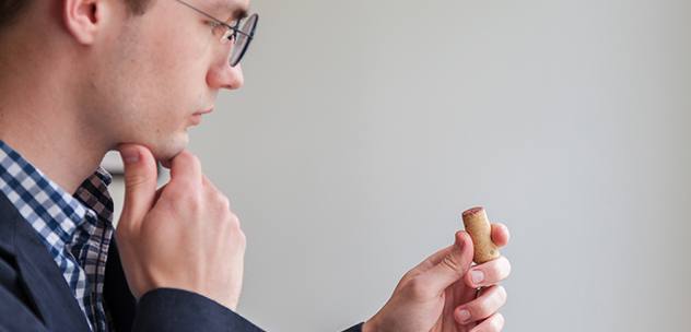 man with glasses and fingers on chin thoughtfully looking at a wine cork in his other hand
