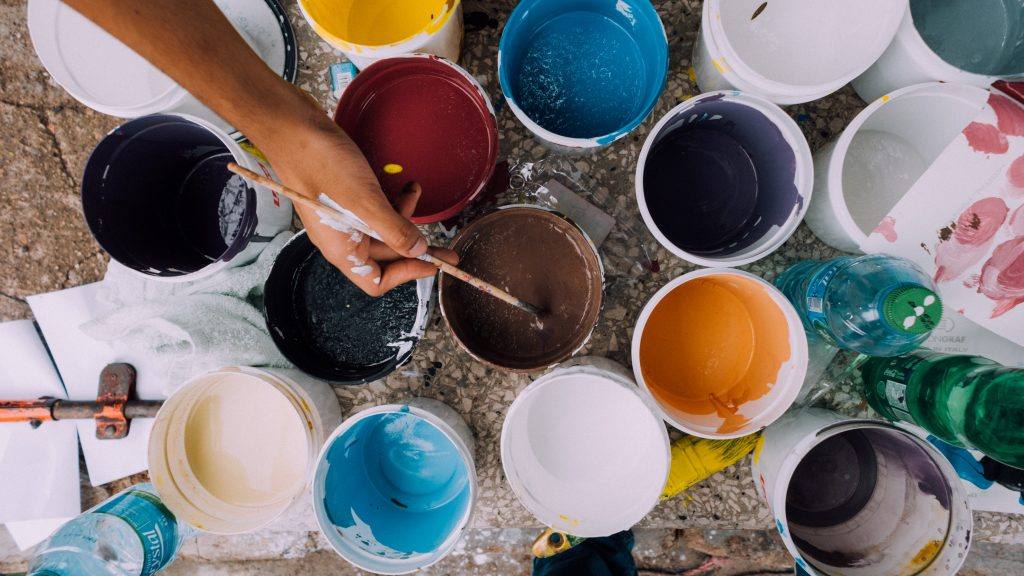 many cans of different colors of paint with one hand holding a paintbrush inside the center one