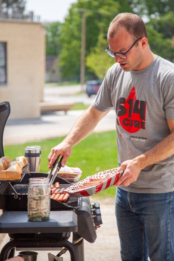 man in short sleeve t-shirt cooks hot dogs at a grill outside