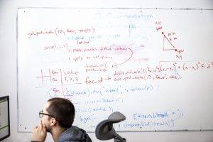 man sits at computer in front of whiteboard filled with mathematical figures