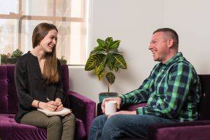 man and woman sit on purple couches and laugh