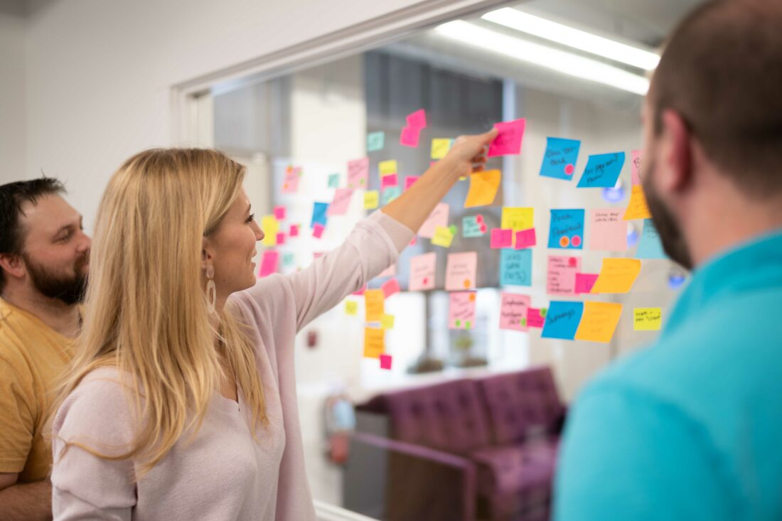 Three coworkers gathered around a wall of post-it notes. girl sticking a note on the wall and contributing to brainstorming session