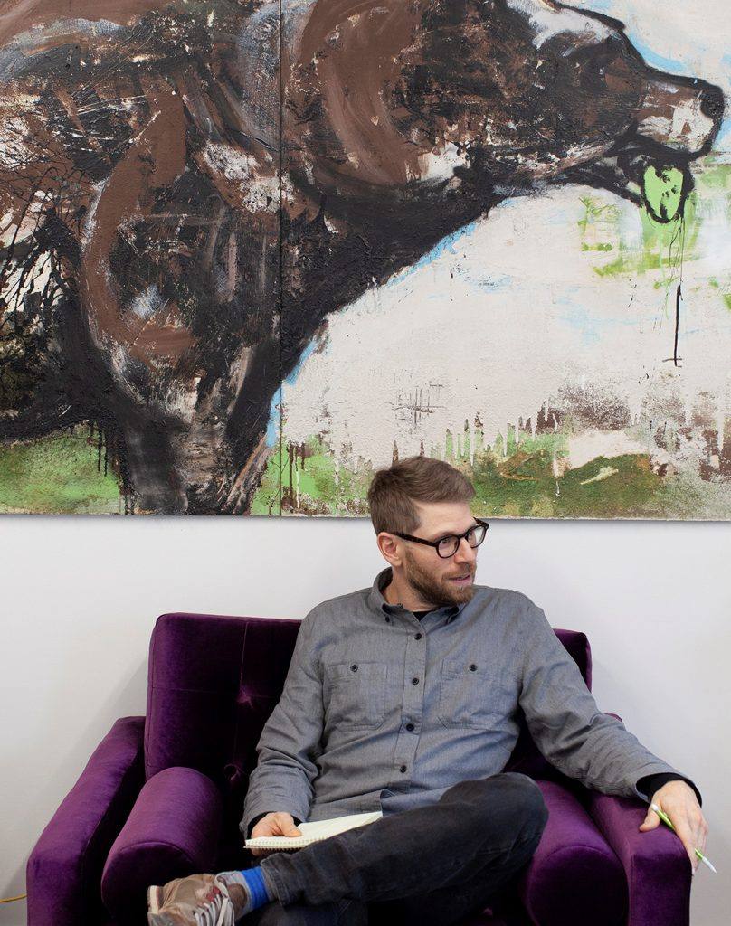 Man sits in an armchair with a notebook in front of a large painting of a bear