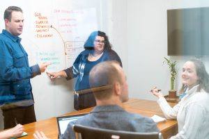 Oneupweb employees discuss market research and write notes on a whiteboard wall