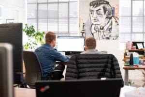 two men in an office sit with their backs to the camera looking at the same computer