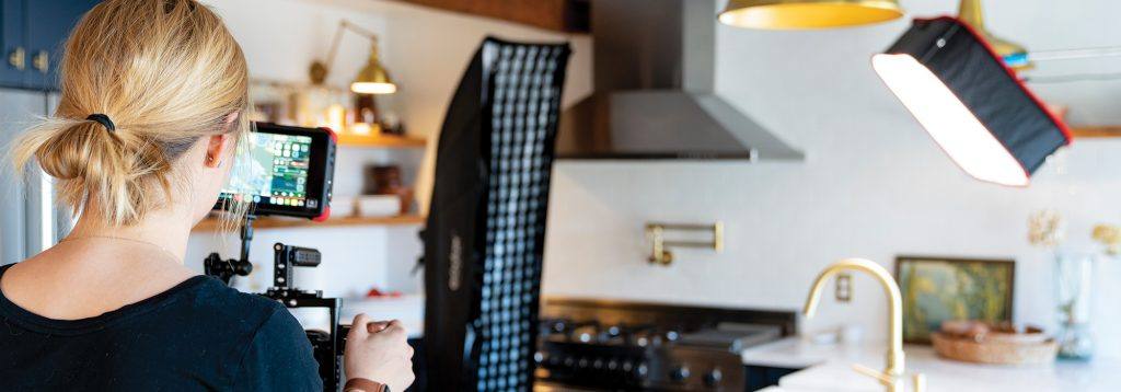 The back of a videographer's head, her camera, and her lighting setup in a nice kitchen