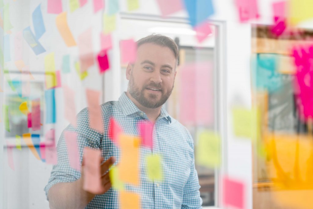 Looking through a window, seeing a man pointing at a wall of post-it notes during a brainstorming session at Oneupweb