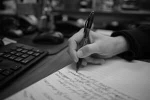 A person's hand writes notes on some notebook paper next to a computer keyboard