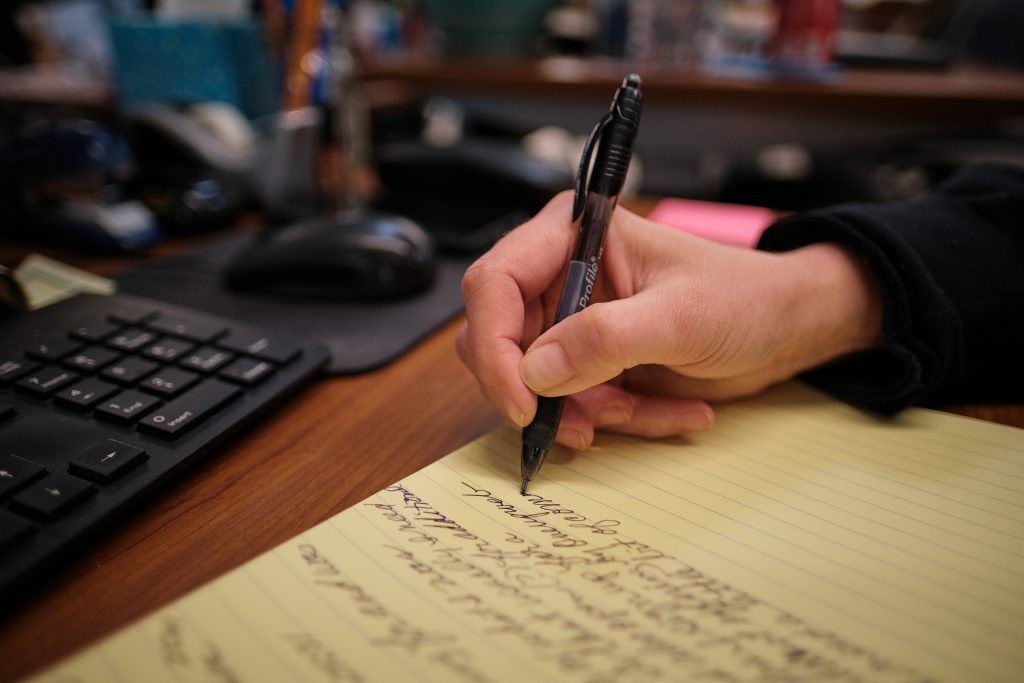 person writing cursive on yellow legal pad notebook next to work desk keyboard