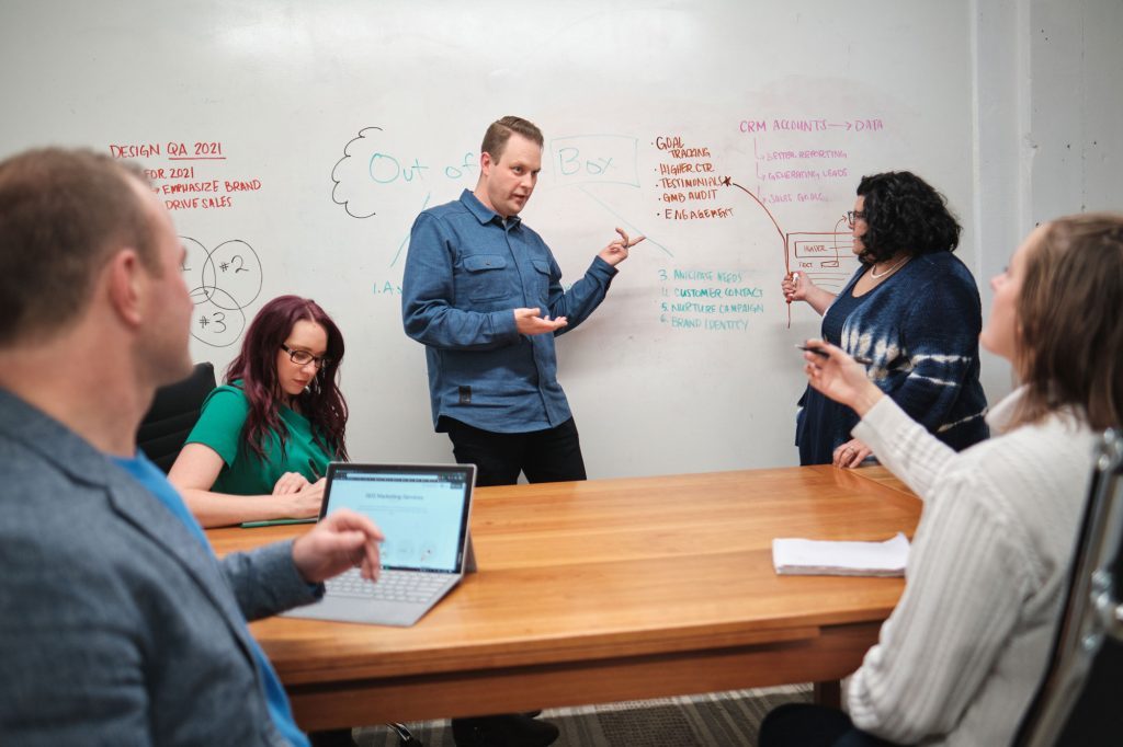 man and woman stand at head of conference room addressing others seated in the room and gesturing at words drawn on whiteboard wall