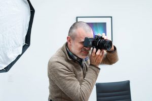 a man holds a digital camera up to his eyes to take a photograph while in an office setting