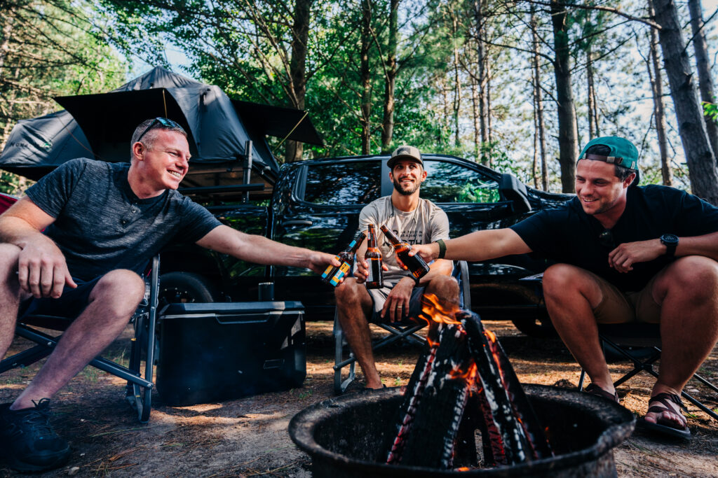 three men sit by a campfire with a truck in the background while clinking beer bottles