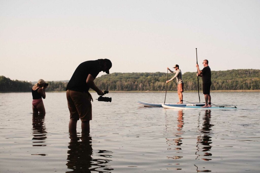 photographers in knee-deep water line up a shoot of paddle boarders on a lake