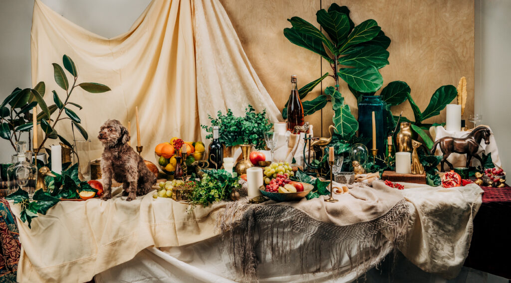 a decadent table of fruit, brass objects, candles, plants and a dog