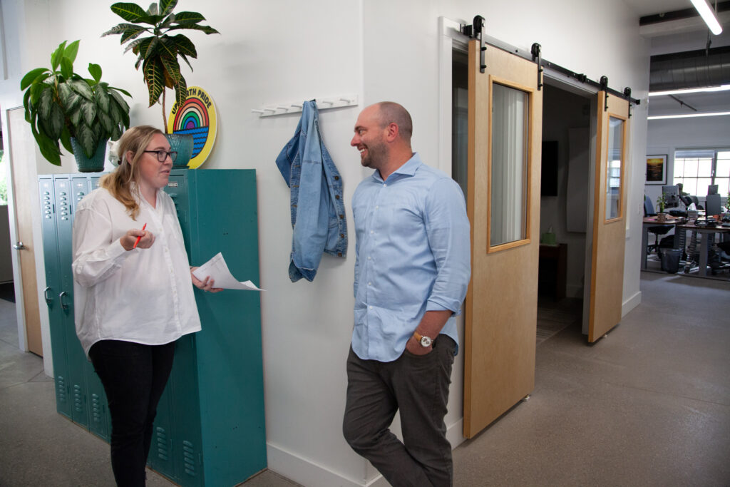 a marketer with blonde hair wearing a white button up and black pants leans against teal lockers holding a pen and notebook while chatting with another marketer wearing a blue button up shirt learning against a hallway wall in the oneupweb office