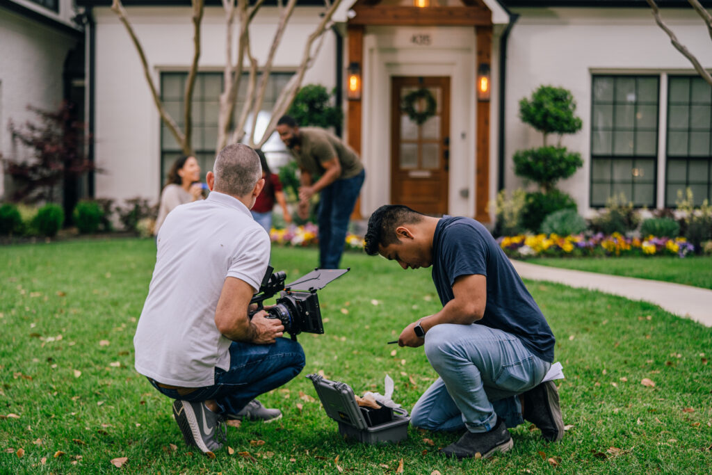 two men crouch over camera equipment in a green grass yard in front of a nice house while filming franchise marketing videos and photography