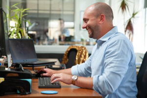 Oneupwebber in a blue shirt, smiling and working at the computer.