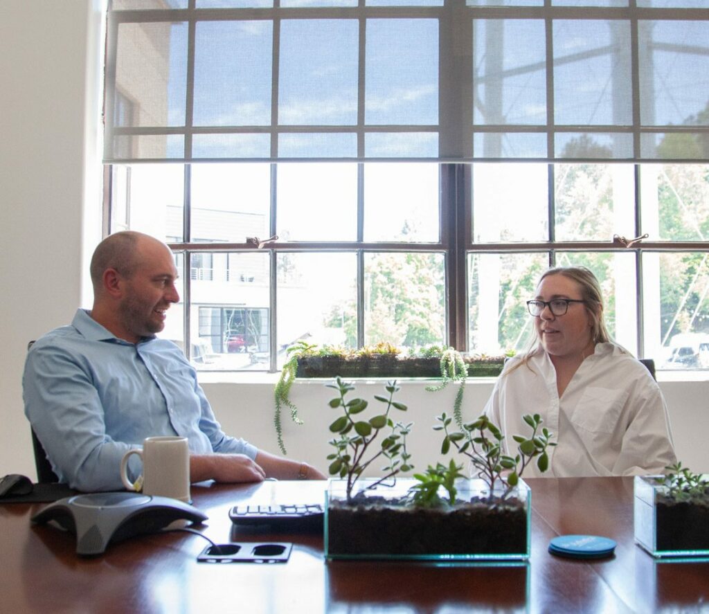 a man in a blue button up shirt and a woman in a white button up talk at a meeting room table with bright windows behind them and plants in front on the table top