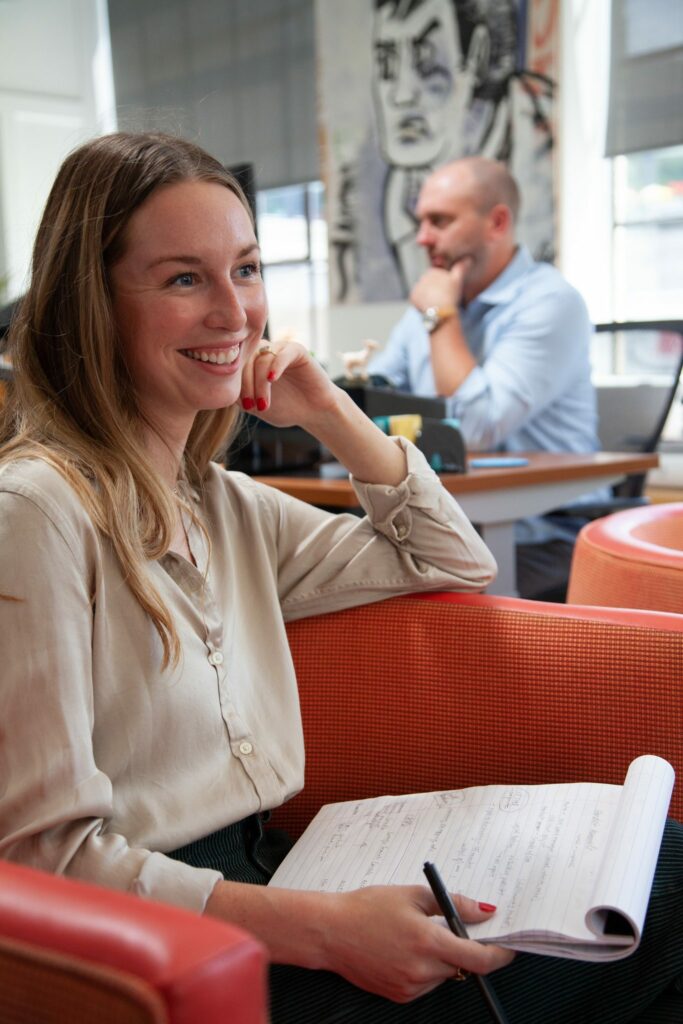 a female marketer sits in a comfy chair while writing in a notebook, smiling to herself, a male coworker, looking at his computer with his hand on his chin appears lost in thought