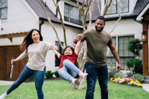 a husband and wife swing their daughter on her arms playing in the front yard of a large, nicely painted house