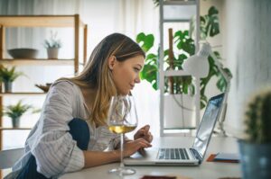 a woman leans over her laptop browsing wineries online while drinking a glass of wine