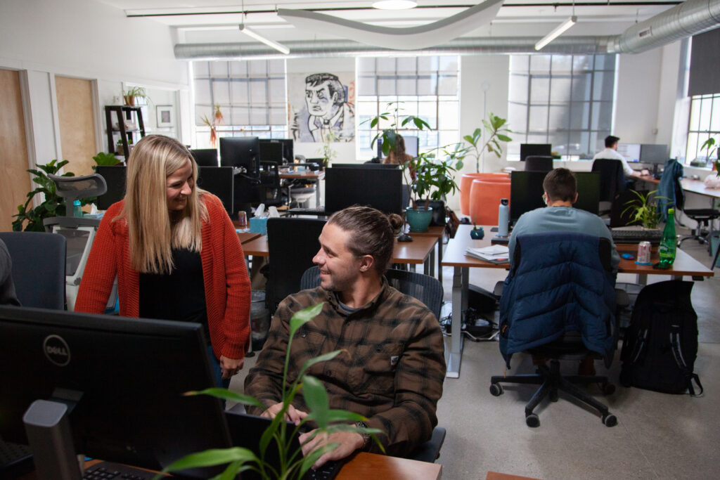 a female marketer stands at a coworkers desk, chatting while smiling with a male coworker sitting at his desk in front of a computer discussing marketing strategy