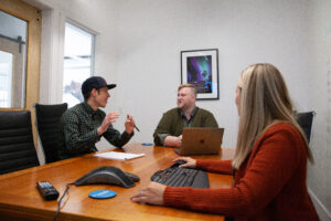 three marketers sit in an office room at a large desk chatting and gesticulating with their hands about a marketing project