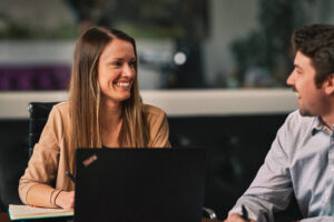 two marketers work side by side on their laptops in a meeting room, discussing recruiting campaigns