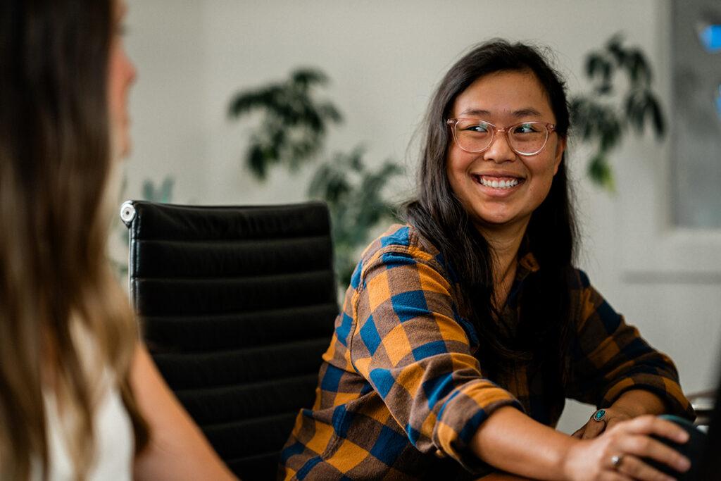 a woman working in an office at a computer smiles while chatting with a colleague